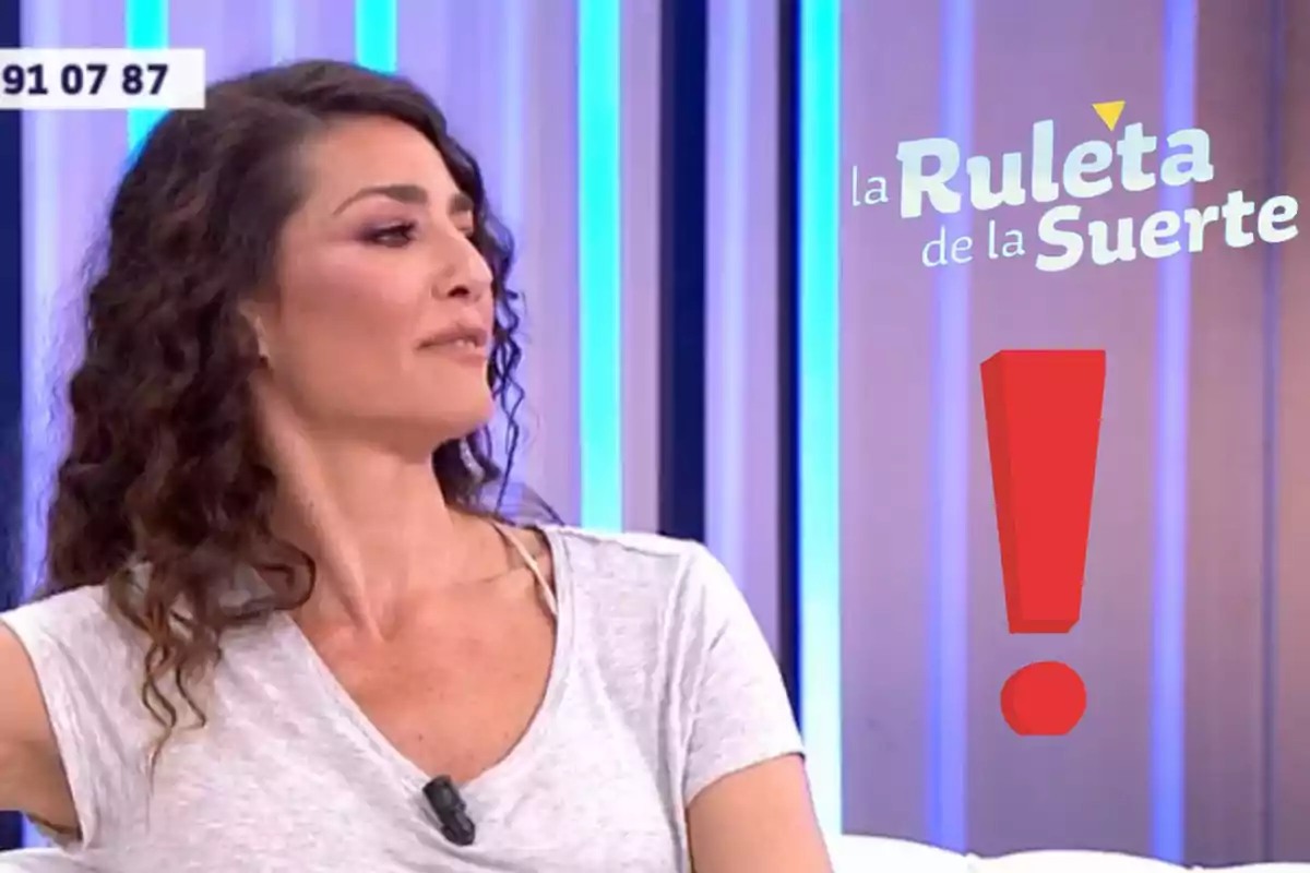 Laura Moure de cabello rizado y camiseta clara sentada en un set de televisión con el logo de La Ruleta de la Suerte y un signo de exclamación rojo a su lado