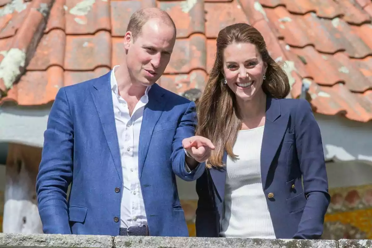 Una pareja sonriente vestida de manera elegante con chaquetas azules posando frente a un techo de tejas. Una pareja sonriente vestida de manera elegante con chaquetas azules posando frente a un techo de tejas.
