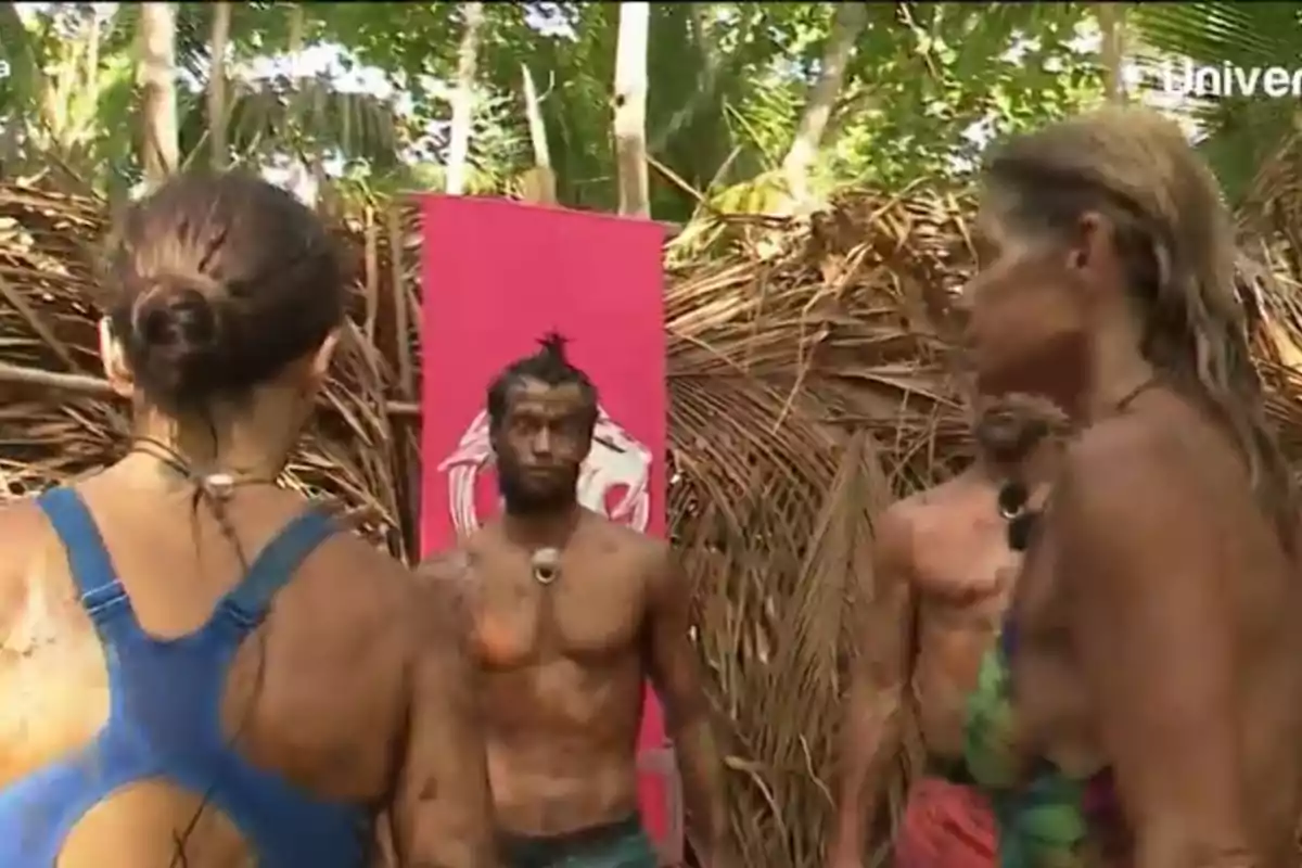 Un grupo de personas cubiertas de barro se encuentra en un entorno selvático, con hojas secas y un cartel rosa de fondo. Un grupo de personas cubiertas de barro se encuentra en un entorno selvático, con hojas secas y un cartel rosa de fondo.