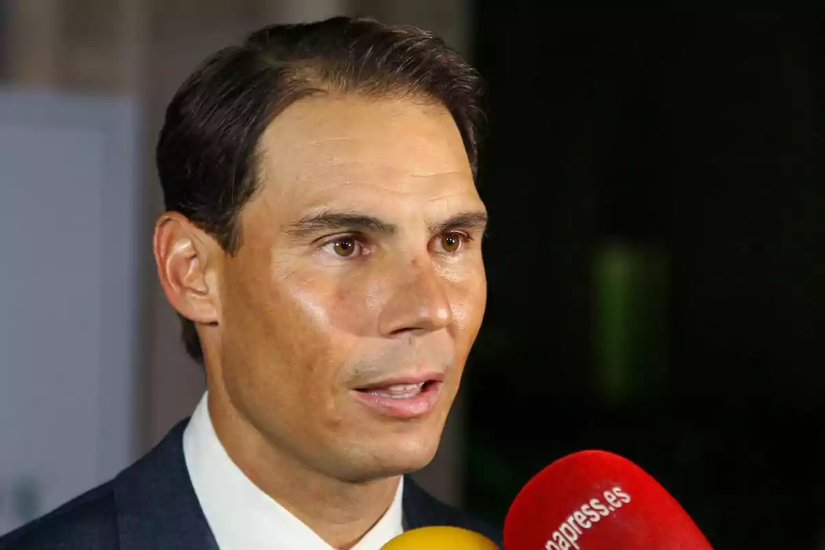 Short, dark-haired man in a suit speaking in front of several colorful microphones Short, dark-haired man in a suit speaking in front of several colorful microphones
