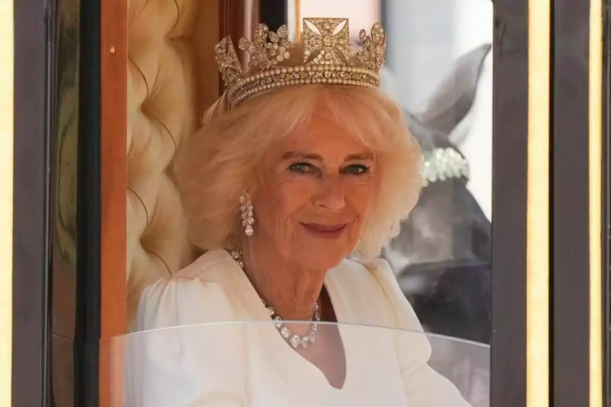Una mujer con cabello rubio y una corona adornada con joyas, vestida de blanco, sonríe desde el interior de un carruaje. Una mujer con cabello rubio y una corona adornada con joyas, vestida de blanco, sonríe desde el interior de un carruaje.