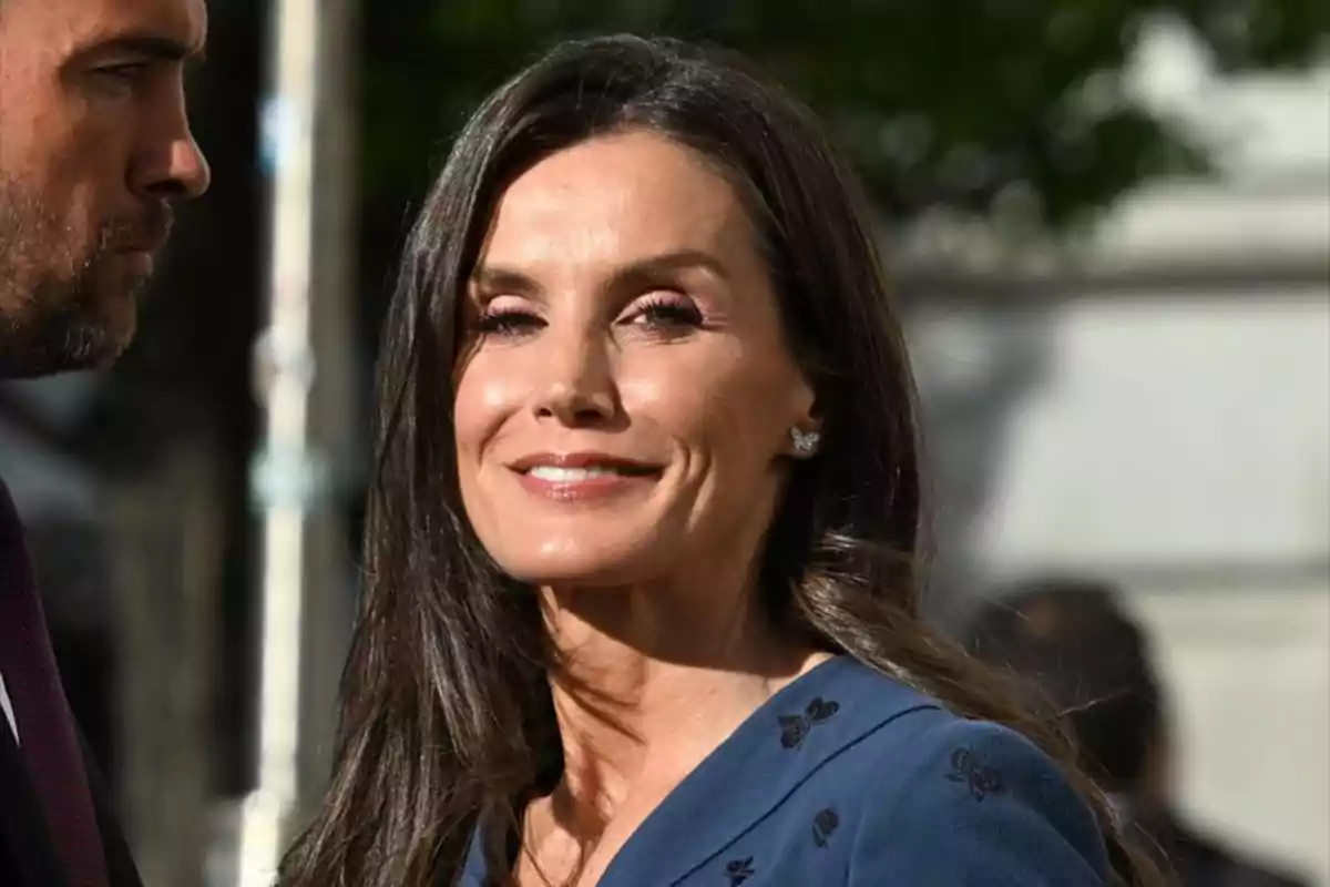 La reina Letizia sonriente con cabello oscuro y largo lleva un vestido azul con detalles bordados y está al aire libre. La reina Letizia sonriente con cabello oscuro y largo lleva un vestido azul con detalles bordados y está al aire libre.