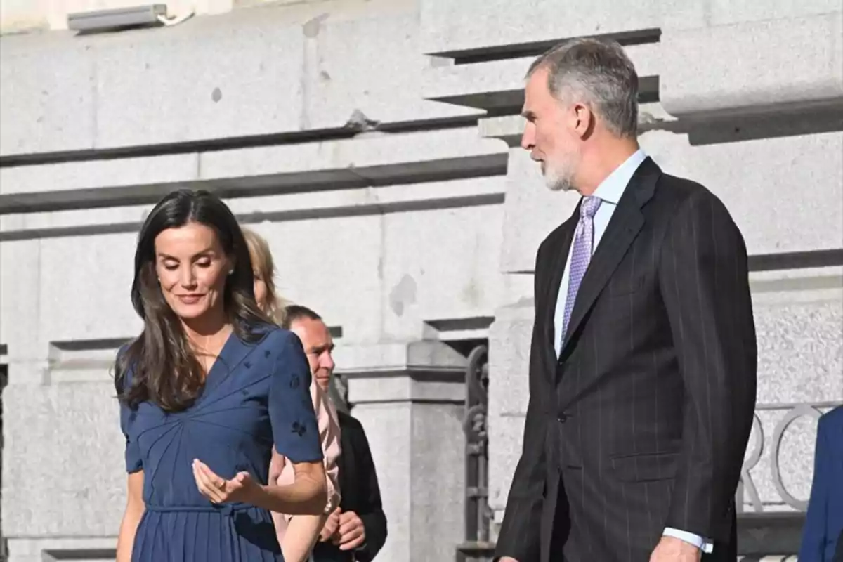 La reina Letizia con un vestido azul y Felipe VI con traje oscuro caminan juntos frente a un edificio de piedra. La reina Letizia con un vestido azul y Felipe VI con traje oscuro caminan juntos frente a un edificio de piedra.