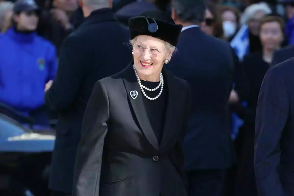 Una mujer mayor con gafas y sombrero negro sonríe mientras lleva un conjunto elegante de color negro con un collar de perlas, rodeada de personas en un evento al aire libre. Una mujer mayor con gafas y sombrero negro sonríe mientras lleva un conjunto elegante de color negro con un collar de perlas, rodeada de personas en un evento al aire libre.