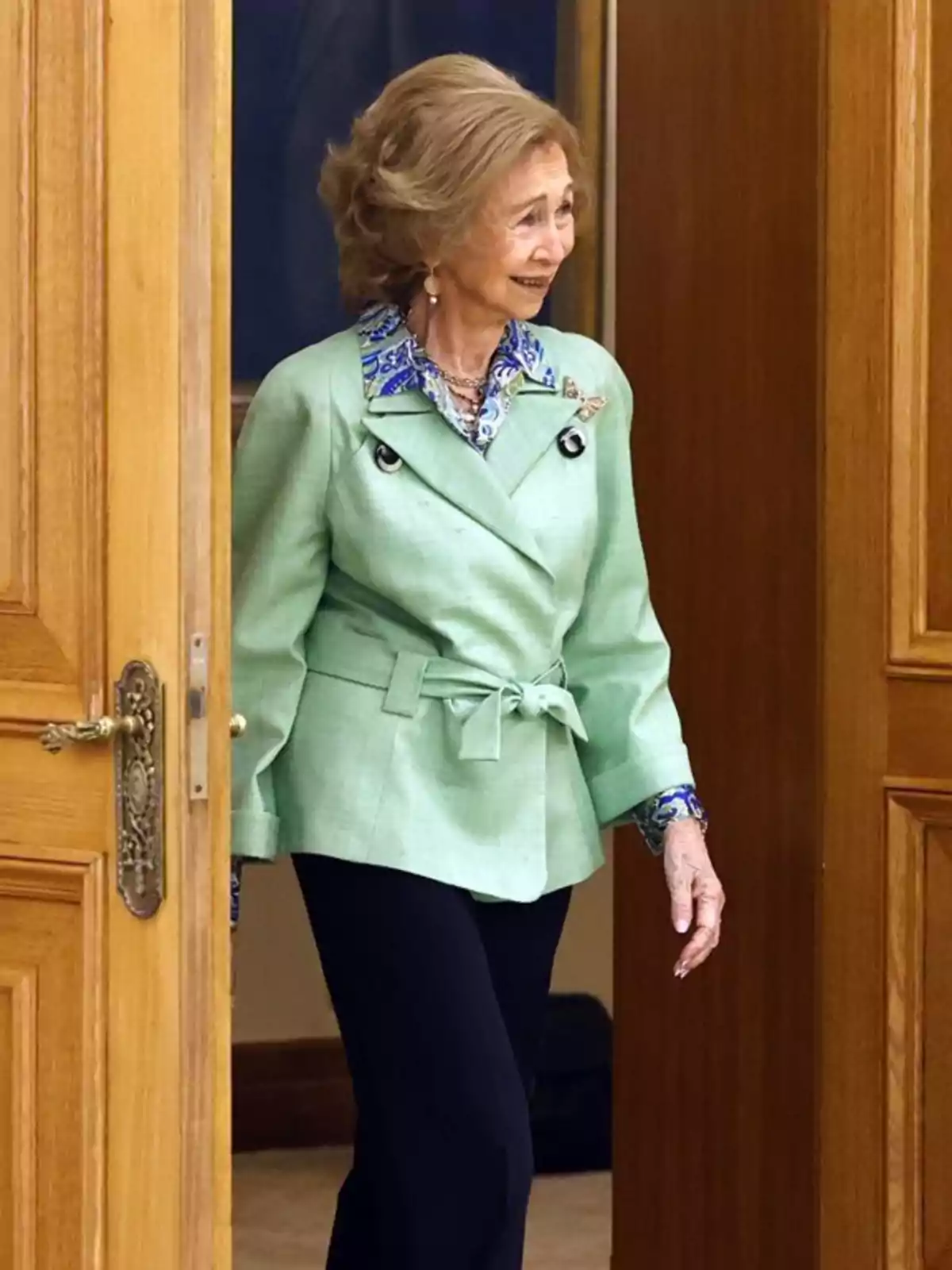 La reina Sofía mayor con chaqueta verde claro y pantalón oscuro entrando por una puerta de madera La reina Sofía mayor con chaqueta verde claro y pantalón oscuro entrando por una puerta de madera