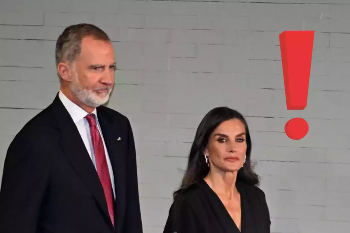 El rey Felipe y la reina Letizia elegantemente vestidos posan frente a una pared gris con un gran signo de exclamación rojo a su lado