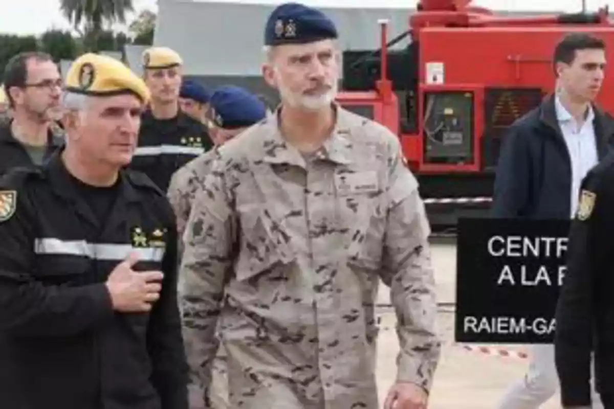 El rey Felipe VI y un grupo de personas con uniformes militares y boinas de diferentes colores camina al aire libre cerca de un edificio y un vehículo rojo. El rey Felipe VI y un grupo de personas con uniformes militares y boinas de diferentes colores camina al aire libre cerca de un edificio y un vehículo rojo.