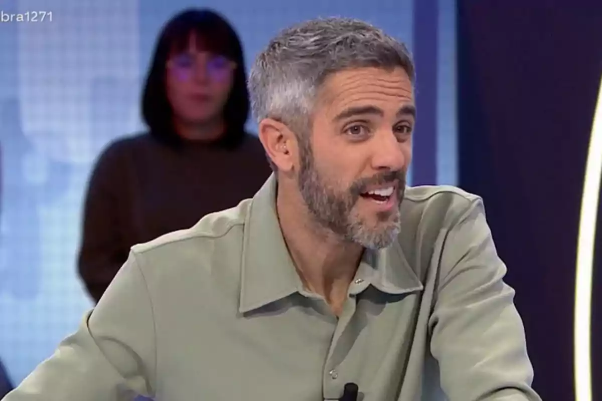 Roberto Leal con barba y cabello canoso sonríe mientras está sentado en un estudio de televisión, con una persona desenfocada al fondo. Roberto Leal con barba y cabello canoso sonríe mientras está sentado en un estudio de televisión, con una persona desenfocada al fondo.