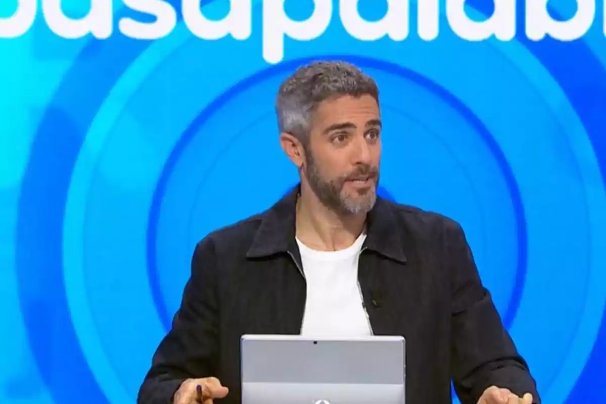 Roberto Leal con barba y cabello canoso está de pie frente a una pantalla azul con letras blancas, sosteniendo un bolígrafo y un dispositivo electrónico. Roberto Leal con barba y cabello canoso está de pie frente a una pantalla azul con letras blancas, sosteniendo un bolígrafo y un dispositivo electrónico.