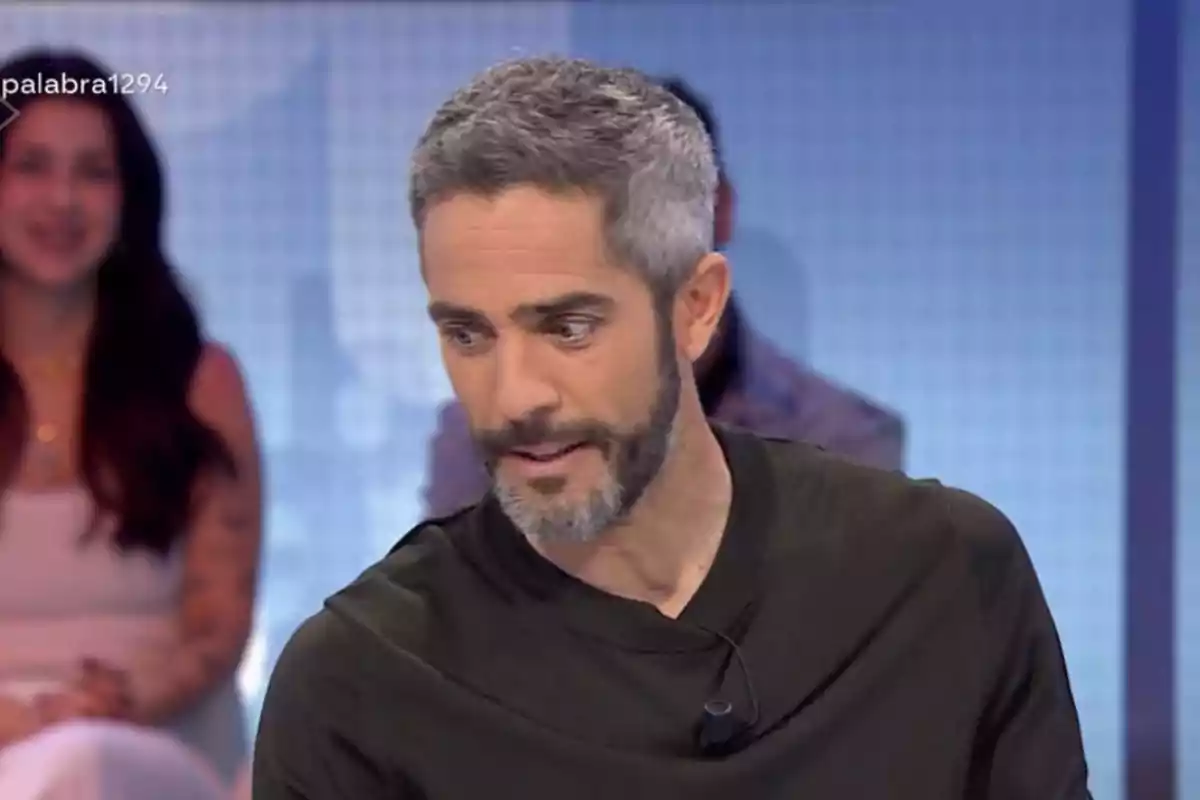 Roberto Leal de cabello y barba canosos con camiseta oscura participando en un programa de televisión con personas desenfocadas al fondo Roberto Leal de cabello y barba canosos con camiseta oscura participando en un programa de televisión con personas desenfocadas al fondo