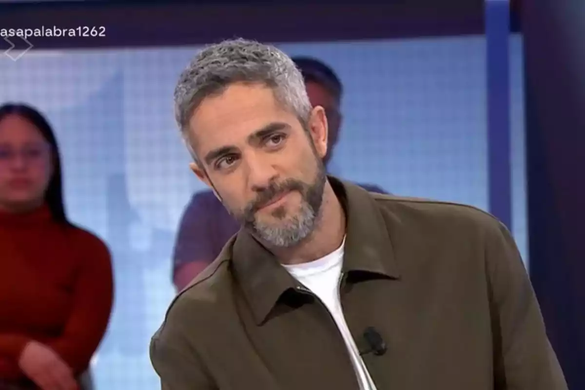 Roberto Leal con barba y cabello canoso está en un estudio de televisión, vistiendo una chaqueta verde y camiseta blanca, con personas desenfocadas al fondo. Roberto Leal con barba y cabello canoso está en un estudio de televisión, vistiendo una chaqueta verde y camiseta blanca, con personas desenfocadas al fondo.