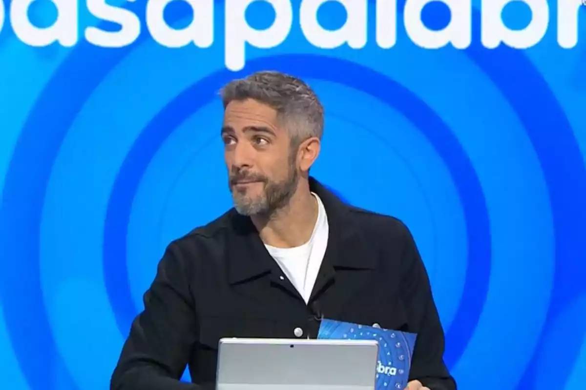 Roberto Leal, presentador de televisión con barba y cabello canoso sostiene una tarjeta frente a un fondo azul con letras blancas. Roberto Leal, presentador de televisión con barba y cabello canoso sostiene una tarjeta frente a un fondo azul con letras blancas.