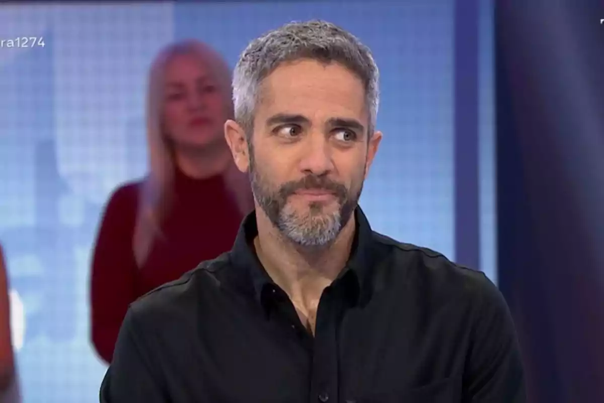 Roberto Leal con barba y cabello canoso está en un estudio de televisión con una expresión pensativa mientras una mujer está desenfocada en el fondo. Roberto Leal con barba y cabello canoso está en un estudio de televisión con una expresión pensativa mientras una mujer está desenfocada en el fondo.