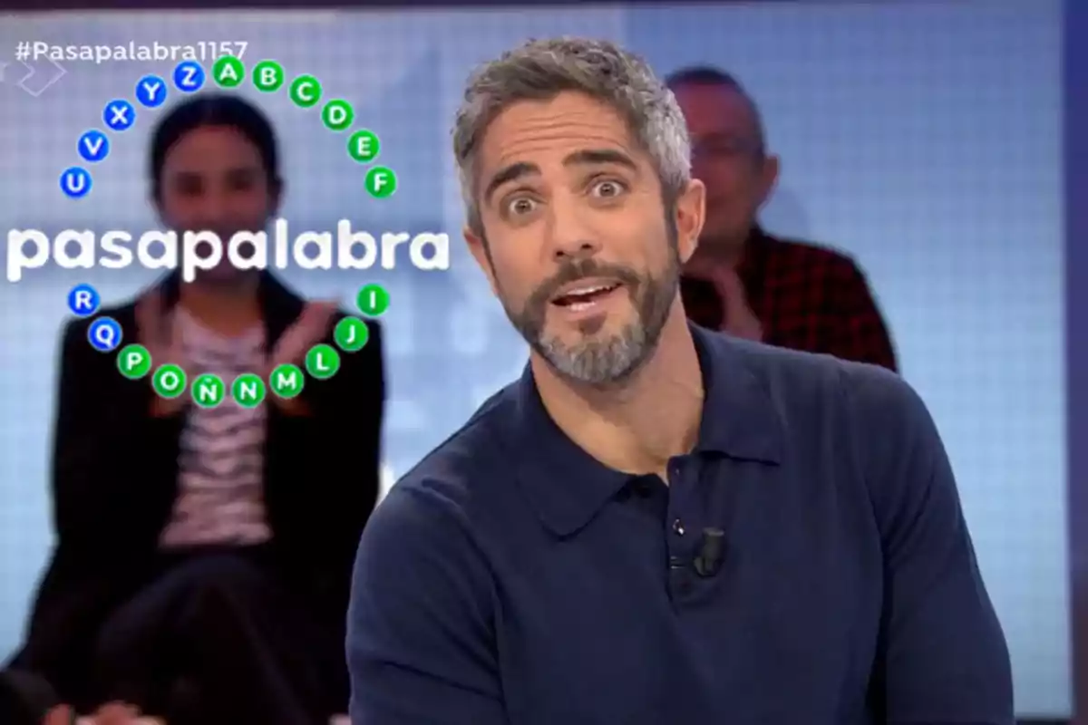 Roberto Leal de televisión con barba y cabello canoso está en un set de concurso con un gráfico circular de letras en el fondo que dice "pasapalabra".