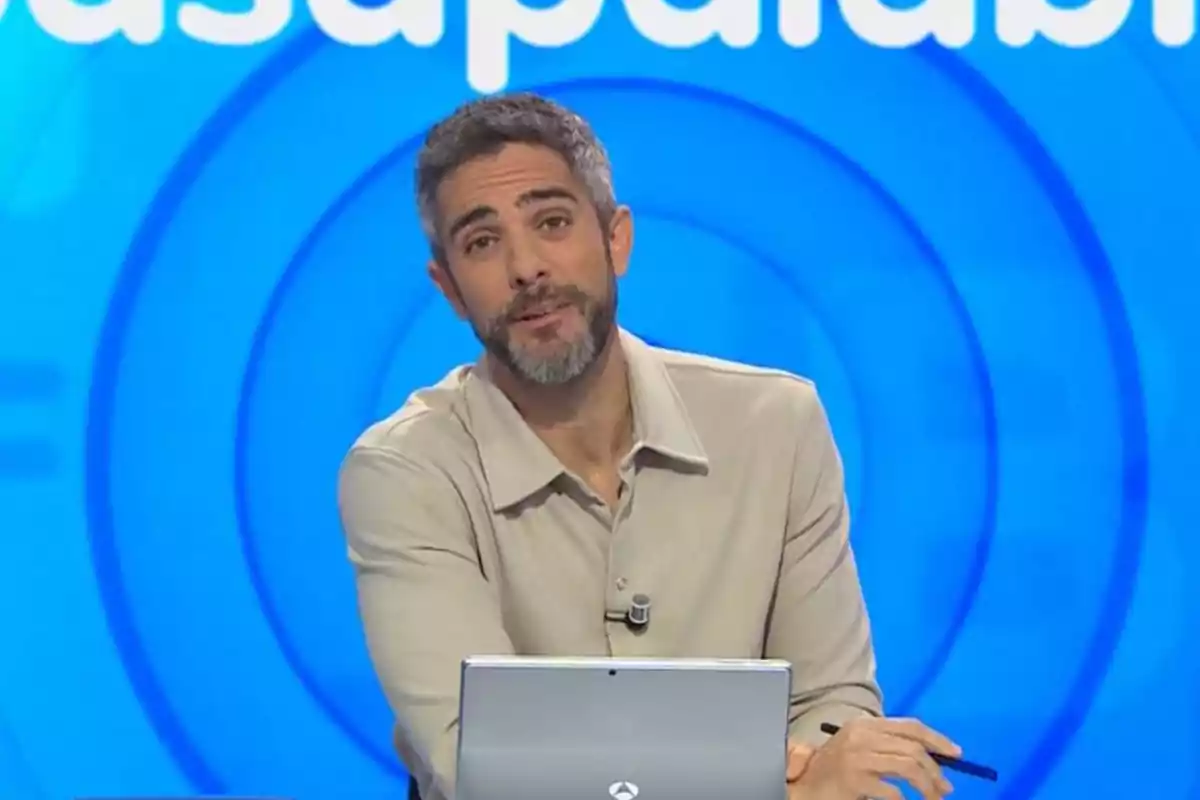 Roberto Leal con barba y cabello corto está sentado frente a una computadora portátil en un set con fondo azul. Roberto Leal con barba y cabello corto está sentado frente a una computadora portátil en un set con fondo azul.