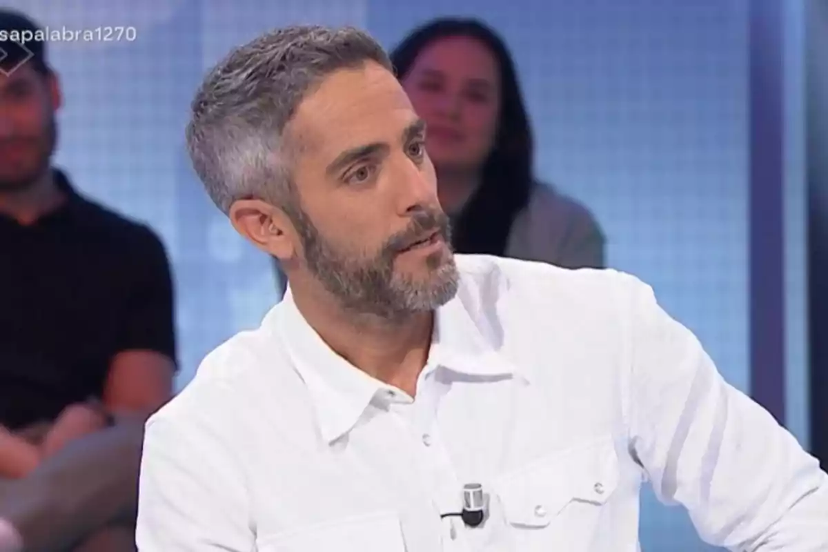 Roberto Leal con barba y cabello canoso lleva una camisa blanca mientras está sentado en un estudio de televisión con personas desenfocadas al fondo. Roberto Leal con barba y cabello canoso lleva una camisa blanca mientras está sentado en un estudio de televisión con personas desenfocadas al fondo.