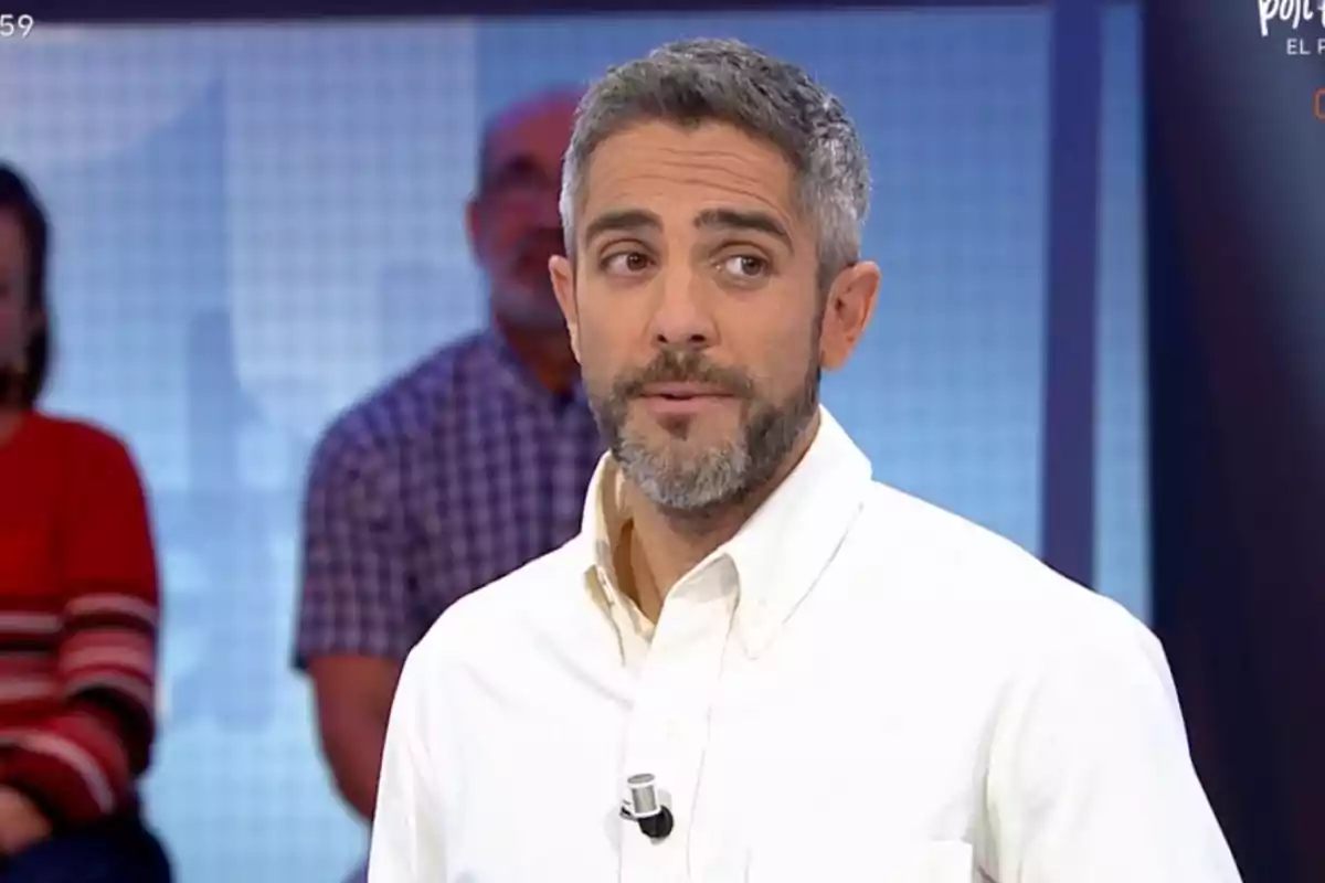 Roberto Leal con barba y cabello canoso, vestido con camisa blanca, está en un estudio de televisión con personas desenfocadas al fondo. Roberto Leal con barba y cabello canoso, vestido con camisa blanca, está en un estudio de televisión con personas desenfocadas al fondo.