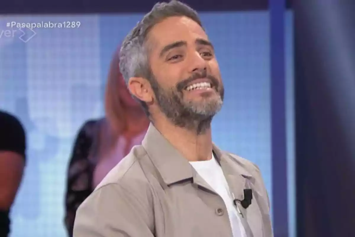 Roberto Leal de cabello corto y barba sonríe en un set de televisión con fondo azul y personas desenfocadas detrás Roberto Leal de cabello corto y barba sonríe en un set de televisión con fondo azul y personas desenfocadas detrás
