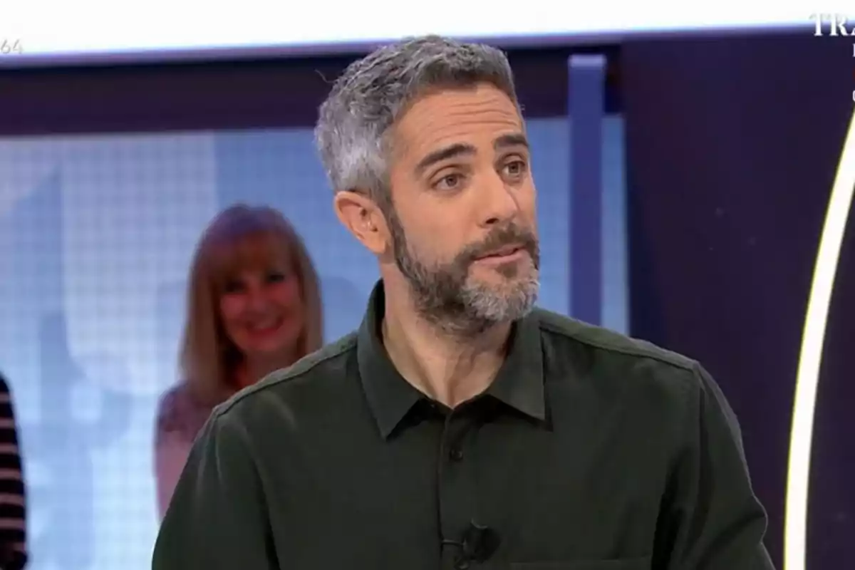 Roberto Leal con barba y cabello canoso está en un estudio de televisión, vistiendo una camisa oscura, mientras una mujer sonríe en el fondo. Roberto Leal con barba y cabello canoso está en un estudio de televisión, vistiendo una camisa oscura, mientras una mujer sonríe en el fondo.