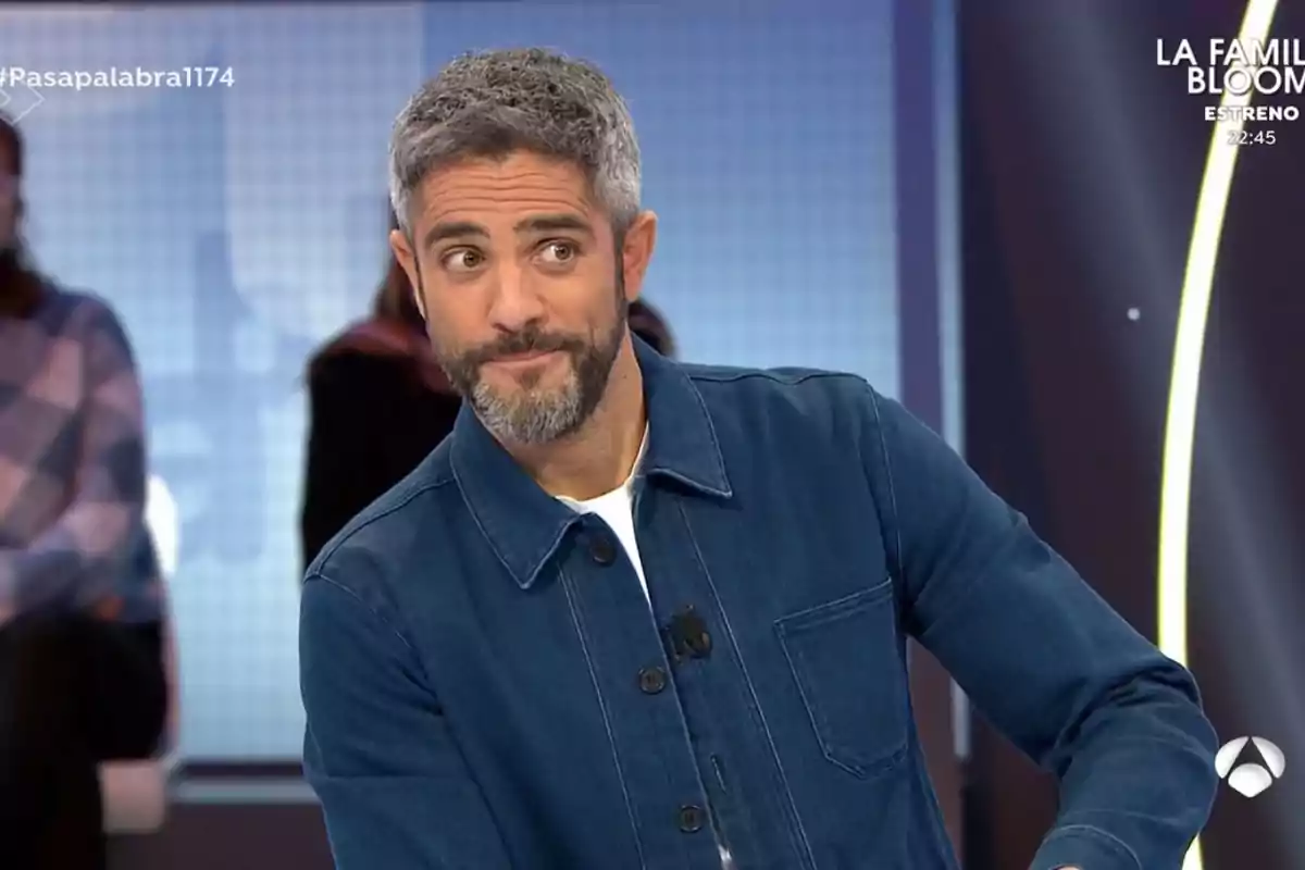 A bearded man with gray hair, wearing a blue jacket, stands on a television set with an out-of-focus background and bright lights.