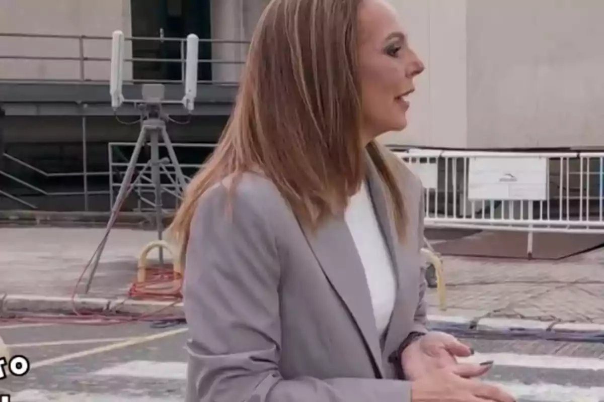 Rocío Carrasco de perfil con cabello largo y lacio lleva un traje gris y está en un entorno urbano con estructuras metálicas y barreras de fondo. Rocío Carrasco de perfil con cabello largo y lacio lleva un traje gris y está en un entorno urbano con estructuras metálicas y barreras de fondo.