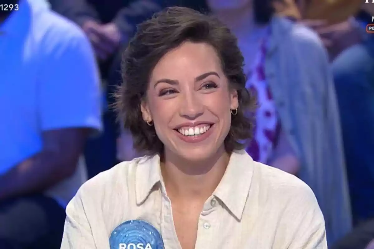 Rosa joven de cabello corto y oscuro sonríe mientras participa en un programa de televisión, lleva una camisa clara y una chapa azul con el nombre Rosa Rosa joven de cabello corto y oscuro sonríe mientras participa en un programa de televisión, lleva una camisa clara y una chapa azul con el nombre Rosa