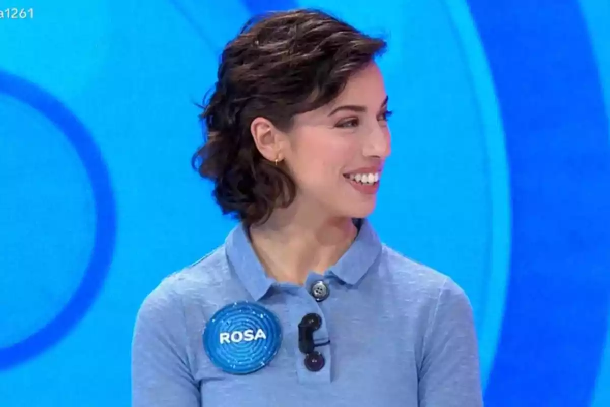 Rosa con cabello corto y oscuro sonríe mientras lleva una camisa azul y un micrófono en un entorno de fondo azul. Rosa con cabello corto y oscuro sonríe mientras lleva una camisa azul y un micrófono en un entorno de fondo azul.