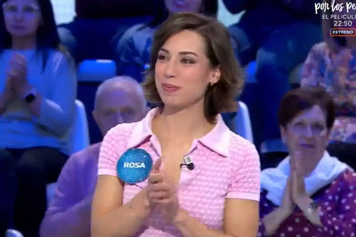 Rosa con una camiseta rosa aplaude mientras está de pie en un estudio de televisión, rodeada de personas en el público. Rosa con una camiseta rosa aplaude mientras está de pie en un estudio de televisión, rodeada de personas en el público.