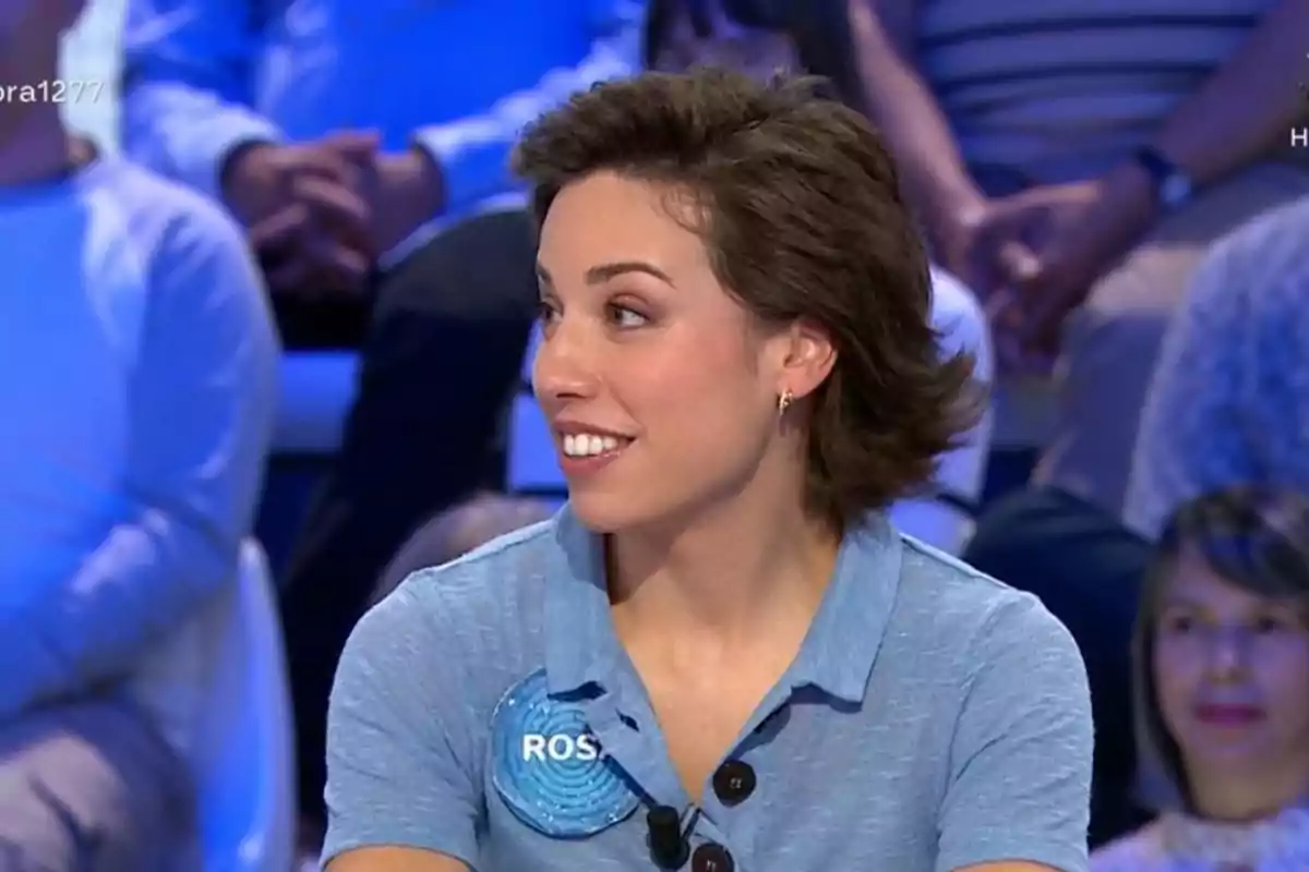 Rosa con cabello corto y castaño, vestida con camisa azul claro y una placa con el nombre Rosa con cabello corto y castaño, vestida con camisa azul claro y una placa con el nombre