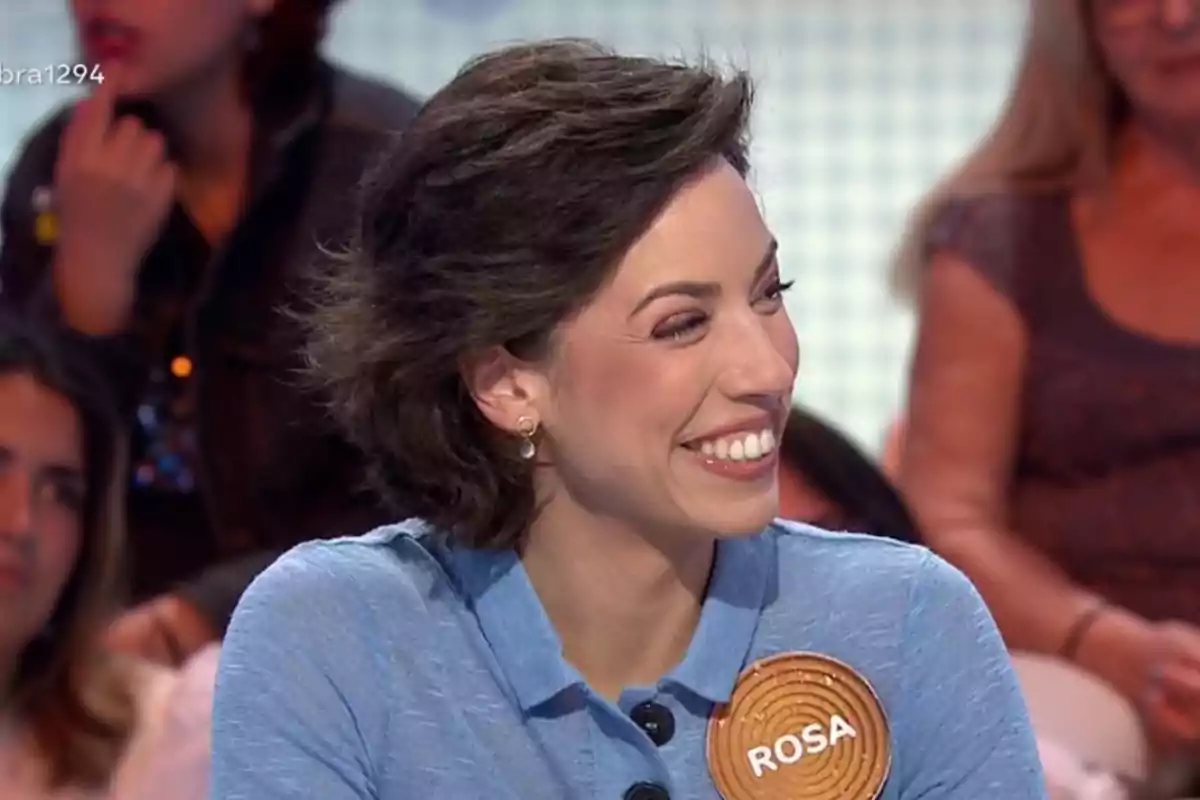 Rosa sonriente con cabello corto y oscuro lleva una camisa azul y un broche con el nombre Rosa en un programa de televisión con público de fondo Rosa sonriente con cabello corto y oscuro lleva una camisa azul y un broche con el nombre Rosa en un programa de televisión con público de fondo