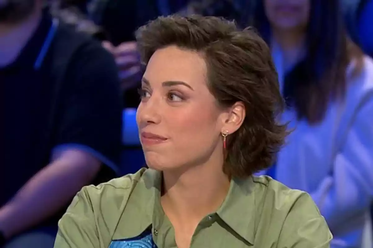 Rosa con cabello corto y oscuro, usando una camisa verde y aretes, está sentada en un entorno de estudio de televisión, mirando hacia un lado. Rosa con cabello corto y oscuro, usando una camisa verde y aretes, está sentada en un entorno de estudio de televisión, mirando hacia un lado.