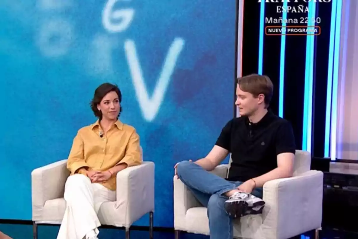 Manu y Rosa sentados en sillones blancos conversan en un set de televisión con fondo azul y letras desenfocadas Manu y Rosa sentados en sillones blancos conversan en un set de televisión con fondo azul y letras desenfocadas