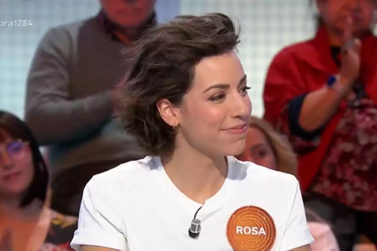 Rosa con cabello corto y oscuro sonríe mientras participa en un programa de televisión, lleva una camiseta blanca con un distintivo que dice Rosa y hay personas en el fondo aplaudiendo. Rosa con cabello corto y oscuro sonríe mientras participa en un programa de televisión, lleva una camiseta blanca con un distintivo que dice Rosa y hay personas en el fondo aplaudiendo.