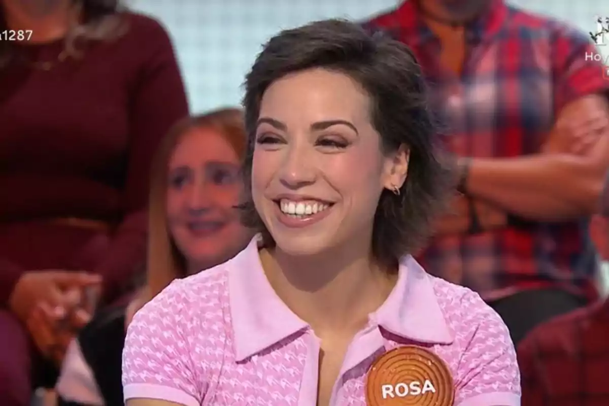 Rosa sonriente con cabello corto y oscuro lleva una camisa rosa y un gafete que dice Rosa mientras participa en un programa de televisión con público al fondo Rosa sonriente con cabello corto y oscuro lleva una camisa rosa y un gafete que dice Rosa mientras participa en un programa de televisión con público al fondo