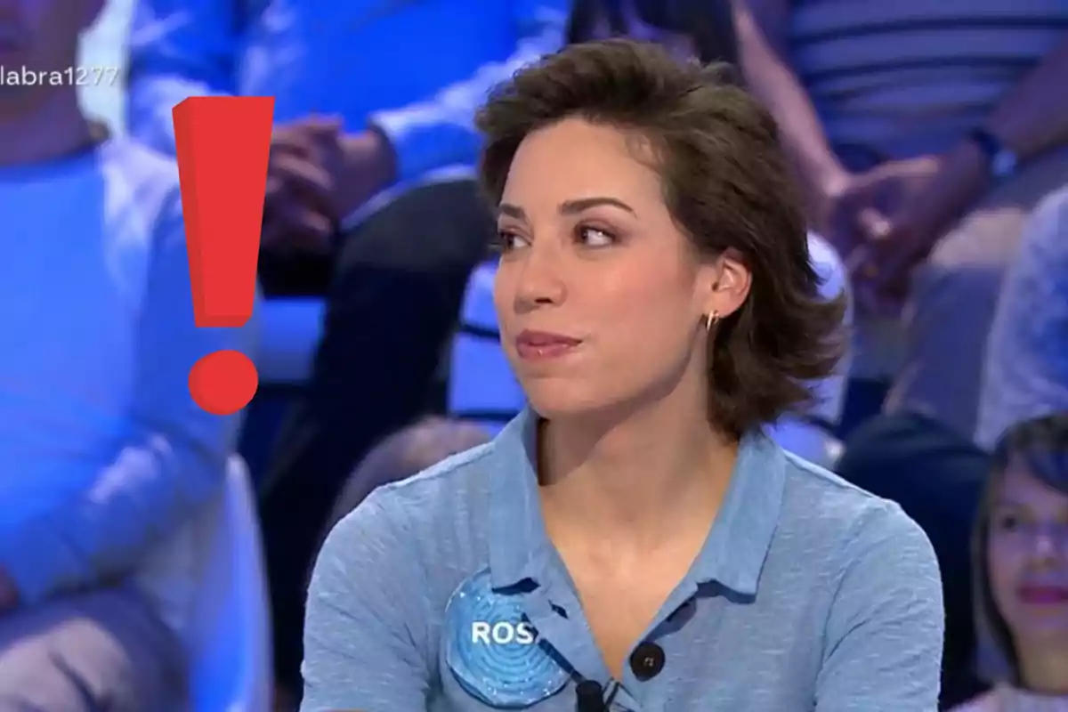Rosa con camisa azul claro sentada en un programa de televisión con un gran signo de exclamación rojo a su lado