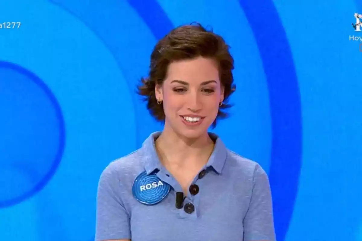 Rosa joven con cabello corto y oscuro, sonriente, vestida con camisa azul claro y un pin que dice Rosa, sobre un fondo azul con círculos concéntricos Rosa joven con cabello corto y oscuro, sonriente, vestida con camisa azul claro y un pin que dice Rosa, sobre un fondo azul con círculos concéntricos