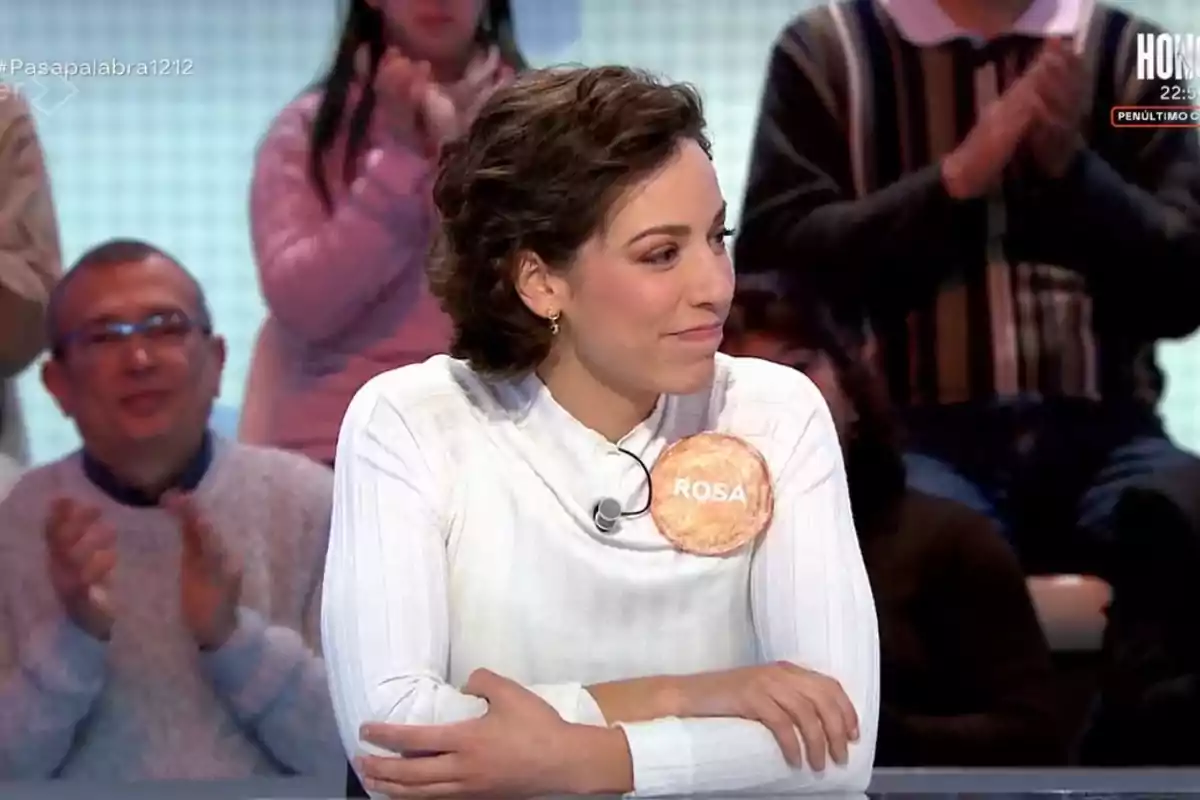 Rosa with short, curly hair, dressed in a white sweater, is sitting on 'Pasapalabra' with an applauding audience in the background.