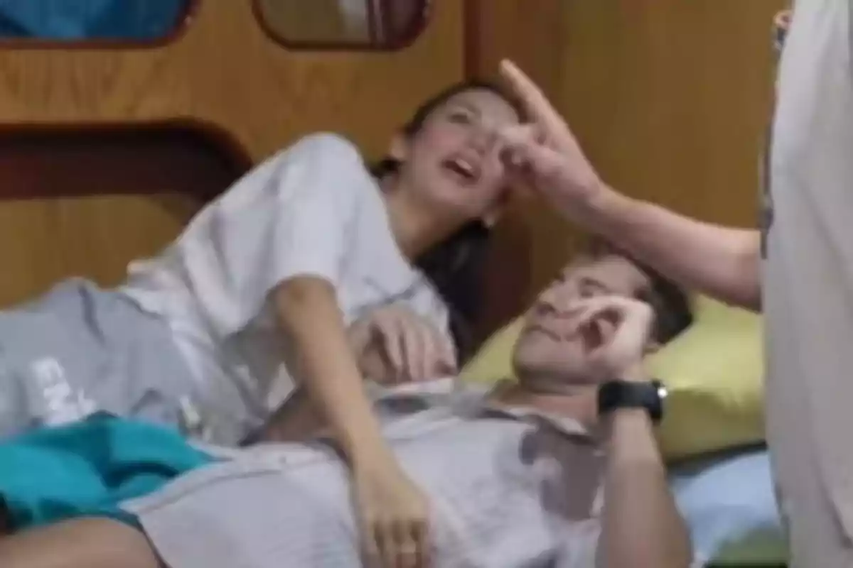 Rosanna Zanetti y David Bisbal están acostados en una cama, mientras una persona de pie señala con el dedo hacia ellos. Rosanna Zanetti y David Bisbal están acostados en una cama, mientras una persona de pie señala con el dedo hacia ellos.