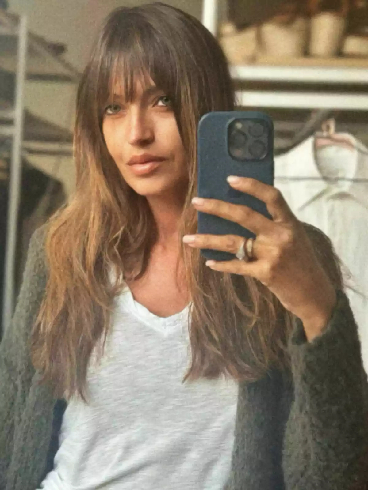 Sara Carbonero con cabello largo y flequillo se toma una selfie frente a un espejo, usando un teléfono móvil con funda azul, lleva una camiseta blanca y un suéter verde, y en el fondo se ven algunas prendas colgadas. Sara Carbonero con cabello largo y flequillo se toma una selfie frente a un espejo, usando un teléfono móvil con funda azul, lleva una camiseta blanca y un suéter verde, y en el fondo se ven algunas prendas colgadas.