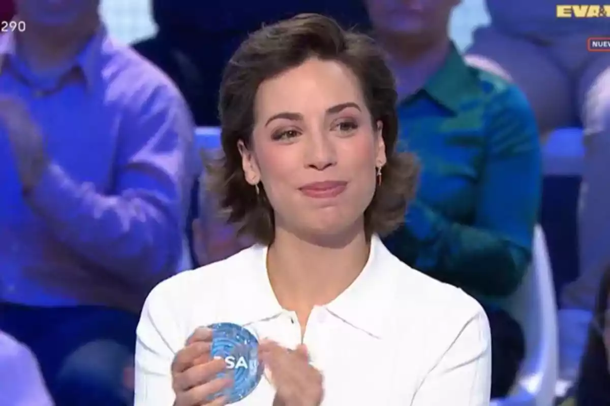 Rosa con cabello corto y camisa blanca sonríe mientras sostiene un objeto redondo azul en un programa de televisión con público de fondo Rosa con cabello corto y camisa blanca sonríe mientras sostiene un objeto redondo azul en un programa de televisión con público de fondo