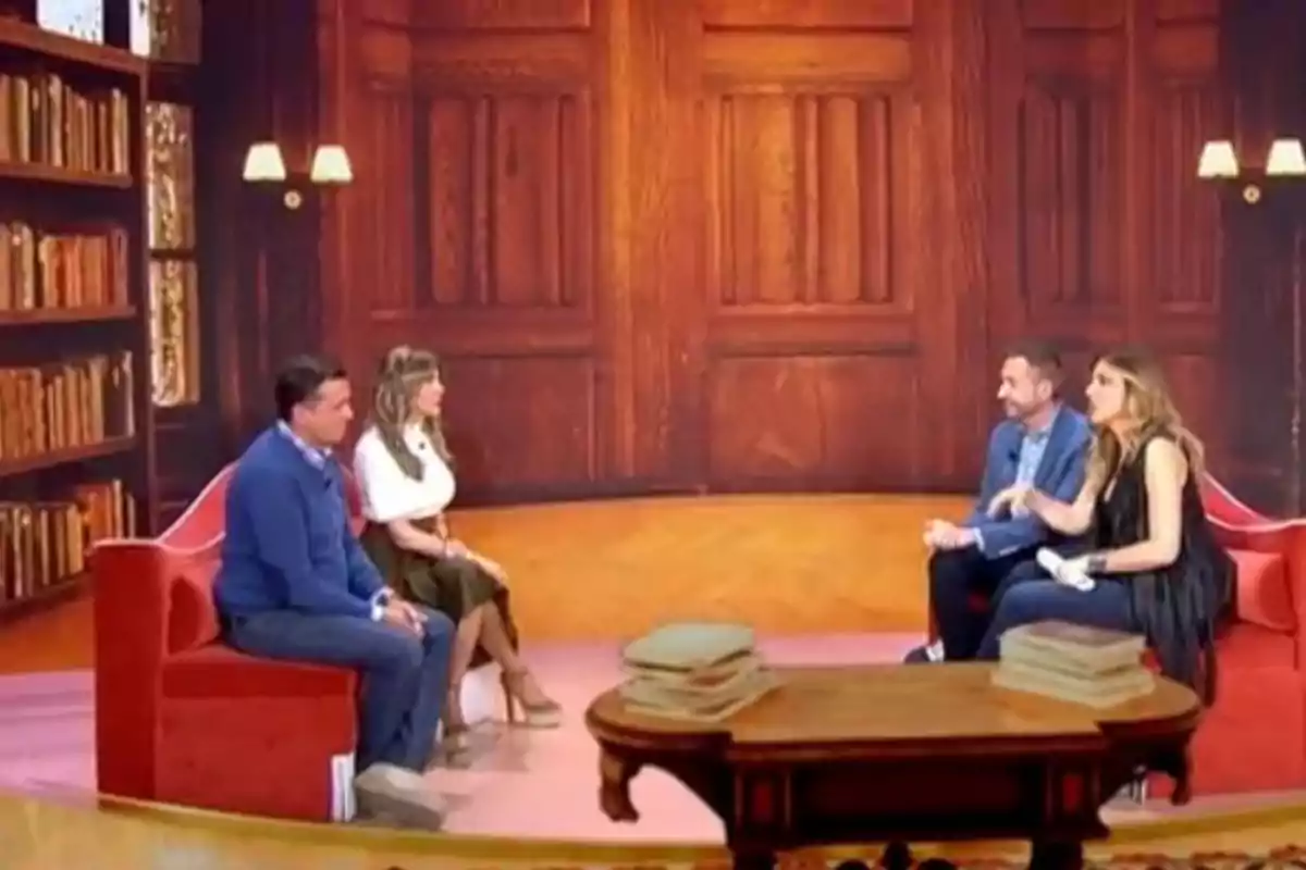 Un grupo de personas está sentado en un set de televisión con una decoración que simula una biblioteca, con estanterías llenas de libros y una mesa en el centro. Un grupo de personas está sentado en un set de televisión con una decoración que simula una biblioteca, con estanterías llenas de libros y una mesa en el centro.