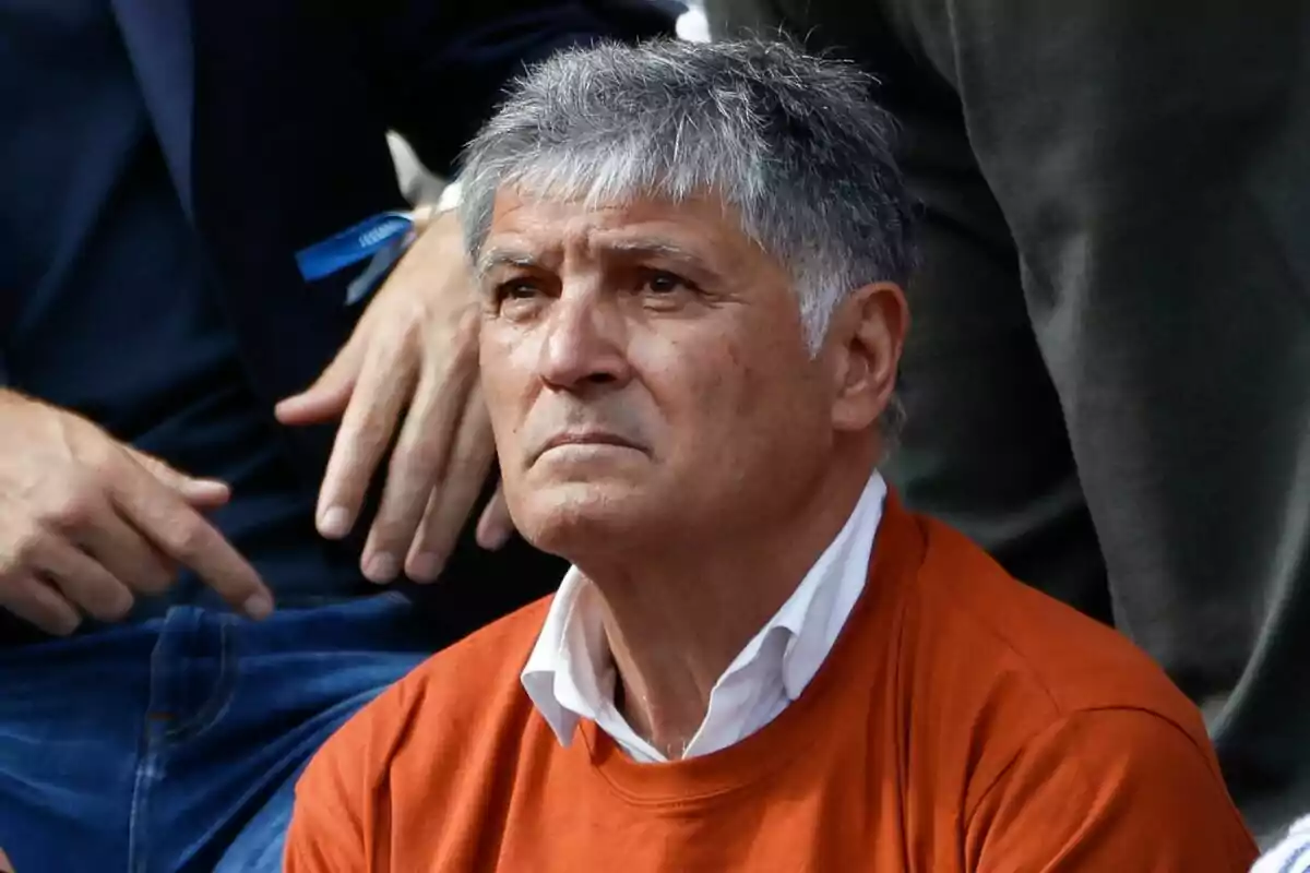Hombre de cabello canoso con expresión seria vistiendo un suéter naranja y camisa blanca rodeado de otras personas Hombre de cabello canoso con expresión seria vistiendo un suéter naranja y camisa blanca rodeado de otras personas