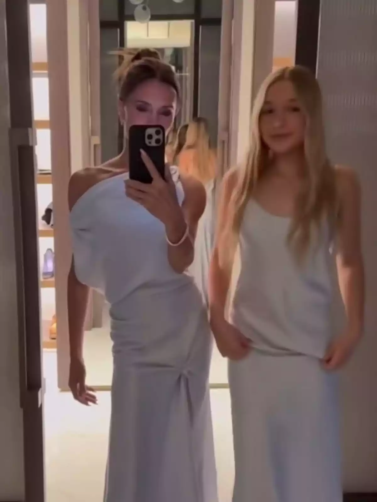 Two people pose in front of a mirror, both dressed in elegant white dresses, while one of them holds a mobile phone to take the picture. Two people pose in front of a mirror, both dressed in elegant white dresses, while one of them holds a mobile phone to take the picture.
