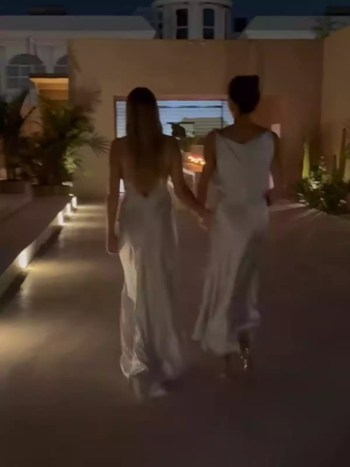 Two people in white dresses walk hand in hand down a brightly lit aisle in an elegant outdoor setting. Two people in white dresses walk hand in hand down a brightly lit aisle in an elegant outdoor setting.