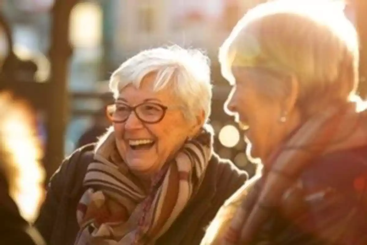 Dos mujeres mayores riendo y disfrutando de una conversación al aire libre con luz de sol iluminando la escena.