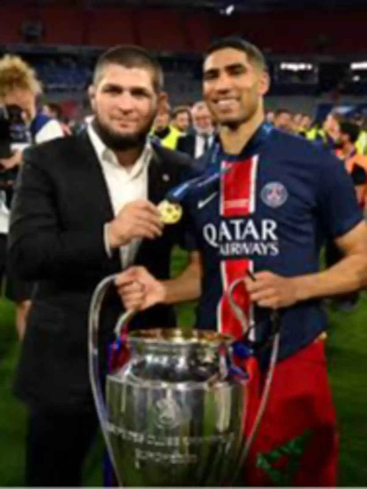 Dos personas sonríen mientras sostienen una medalla y un trofeo de fútbol en un estadio lleno de gente Dos personas sonríen mientras sostienen una medalla y un trofeo de fútbol en un estadio lleno de gente