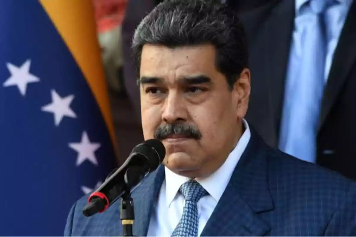 Un hombre con bigote y traje oscuro habla frente a un micrófono con una bandera de fondo.