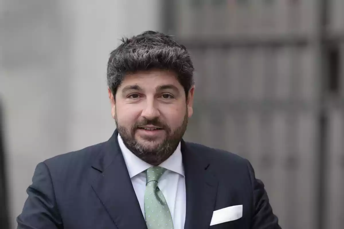 Un hombre con traje oscuro y corbata verde sonríe frente a una estructura de fondo desenfocado.