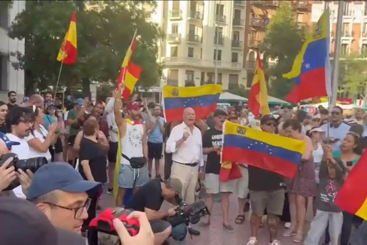 Una multitud de personas se reúne en una plaza, algunas portando banderas de España y Venezuela, mientras varios fotógrafos capturan el momento.