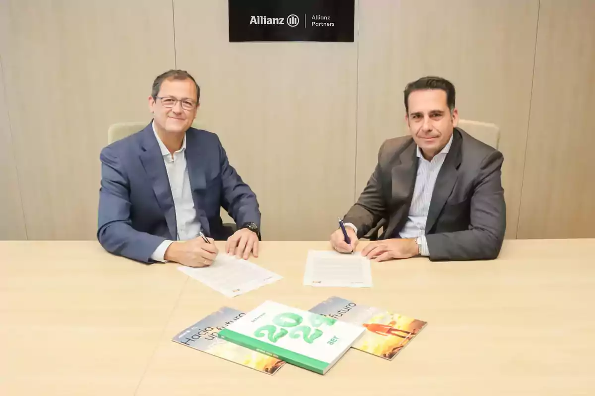 Two men sitting at a table signing documents with books and an Allianz sign on the wall.